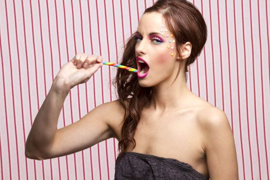 Young woman with colorful makeup and star candy glued to her face, about to bite on a multicolor candy cane