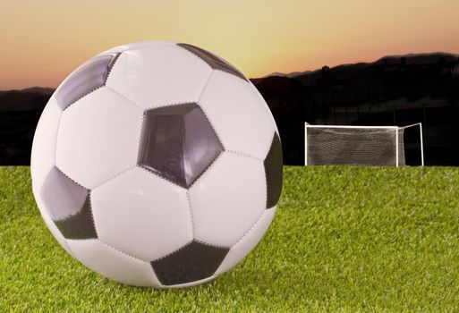 White and black football over grass, with goal and sunset on the back