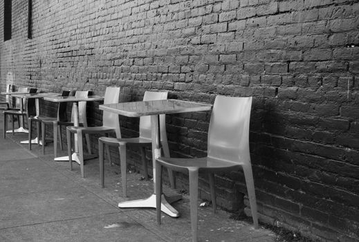 Black and white image of brushed aluminum tables and chairs on a sidwalk against a black brick wall