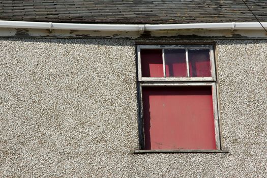 A boarded up window frame in bad repair with missing glass poor guttering a pebble dash wall and slate roof.