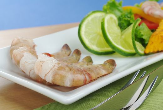 Raw king prawn with lime slices and lettuce on white plate, wooden board and green table mat with cutlery and blue background  (Selective Focus)