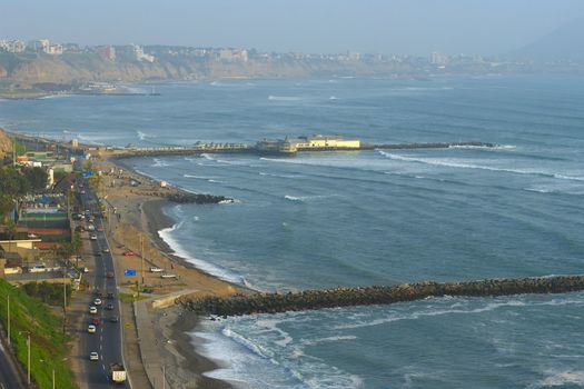 The seaside road in Miraflores and a restaurant built on the pier, with a view on the coastline of Southern Lima in the usual misty weather. The surf breaks are used by many tourists and locals to learn surfing. 
