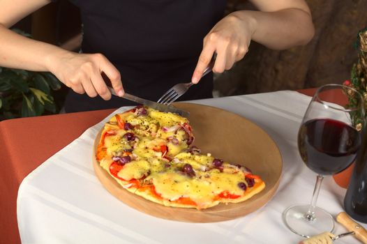 Woman eating vegetarian pizza in a pizzeria with a glass of red wine standing beside the pizza (Selective Focus, Focus on the hands, the cutlery and the pizza around the cutlery)