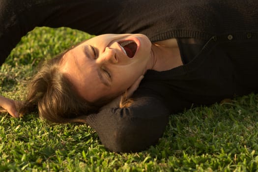 Young Caucasian woman yawning while lying on grass in a park lit by the evening light (Selective Focus, Focus on the eyes)