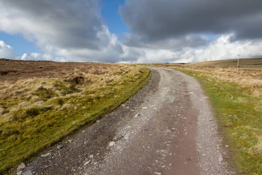 A rough track cuts through moorland towards the horizon with a cloudy blue sky in the distance.