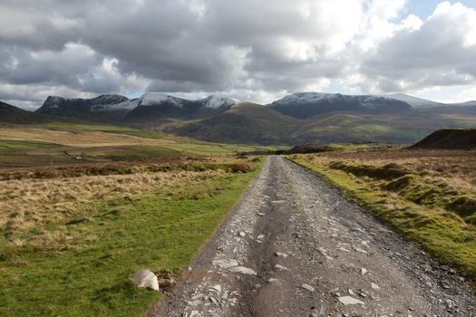 A rough track cuts through moorland towards a cloudy Nantlle ridge in the Snowdonia national park, Wales, UK.