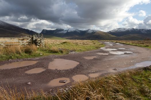 A rough track with water filled potholes cuts through moorland towards a cloudy Nantlle ridge in the Snowdonia national park, Wales, UK.