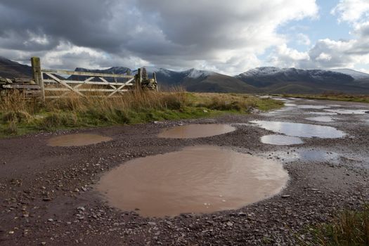 A rough track with water filled potholes cuts through moorland towards a cloudy Nantlle ridge in the Snowdonia national park, Wales, UK.