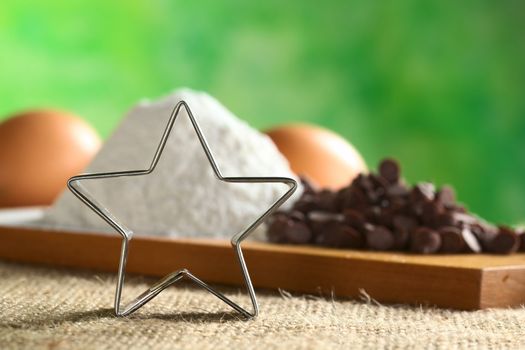 Star-shaped cookie cutter with baking ingredients such as wheat flour, chocolate chips and eggs in the back (Selective Focus, Focus on the cookie cutter)