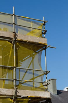 A scaffold structure made from planks and metal bars with yellow safety netting against a clear blue sky.