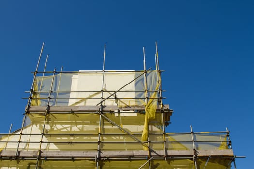 A scaffold structure made from bars and planks with a yellow safety netting around a beige chimney against a clear blue sky.