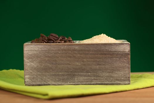 Roasted coffee beans and brown sugar in wooden container on green table mat with dark green background (Selective Focus, Focus on the top of the two piles)