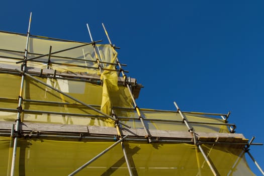 A structure built from scaffolding, bars, planks and clamps with a yellow safety netting against a clear blue sky.