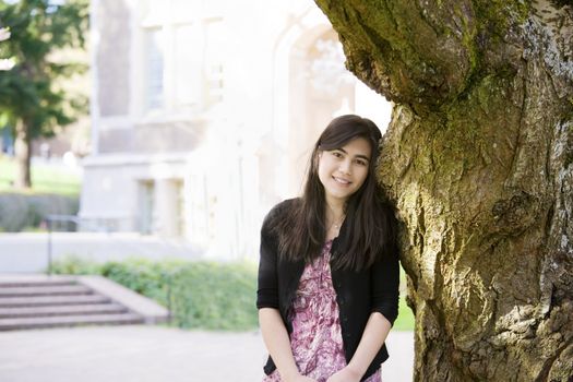 Happy biracial teen girl or young woman leaning against trunk of large tree, smiling. Peaceful scene.