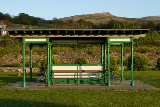 A park pavillion structure with posts, roof and benches on green grass with a hillside against a blue sky in the background.