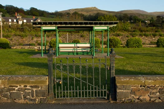 A locked metal gate in a wall leads to an open air pavillion with benches on a grassed area with mountains and blue sky in the background.