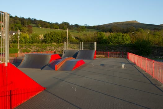 An outdoor skatepark with variuos ramps made from metal painted red with green fields in the background.