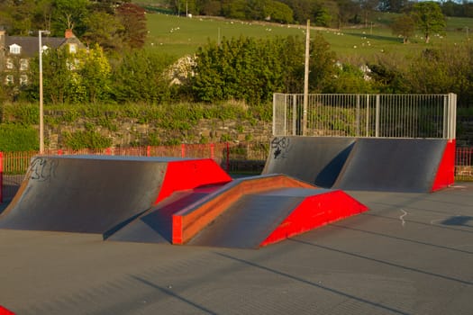 An outdoor skatepark with variuos ramps made from metal painted red with green fields in the background.