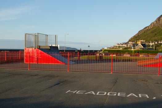 An open air skatepark with a red painted ramp and railings and a sign painted on the floor reading 'HEADGEAR'.