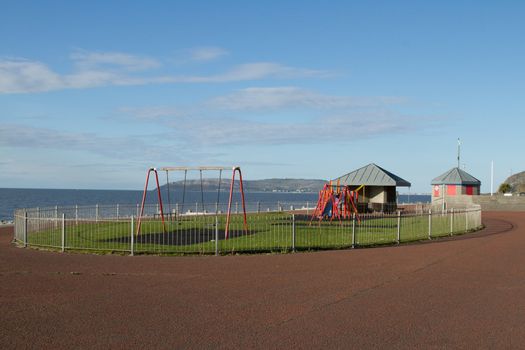 A children's play area with swings and a slide on green grass in an enclosed zone with a metal fence and footpath surround with the sea and an island under a blue cloudy sky in the background.