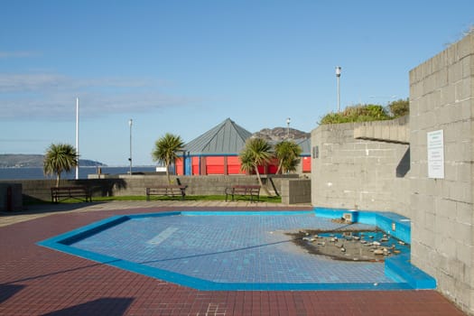 An unused childrens paddling pool, blue and red tiled, in an enclosed area with benches and trees.