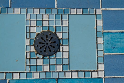 A round metal drain cap with screw fixings set into a blue and white ceramic tile pattern.