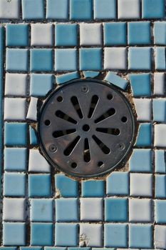 An old round metal drain cap with screw fixings set into a blue and white ceramic tile pattern.