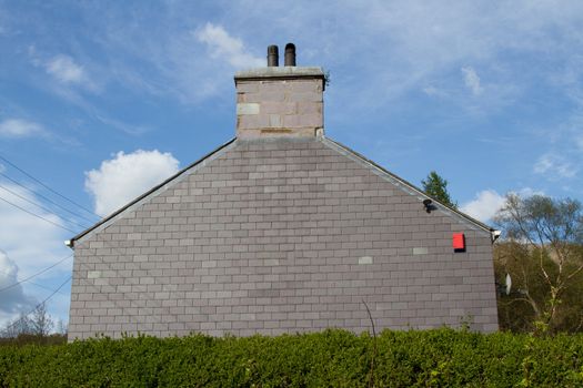 The gable end of a house with chimney and pots with a wall constructed of patterned slate with an alarm and green hedge with a blue cloudy sky in the background.