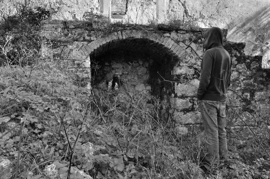 Overgrown plants and hooded man next to arched recess of rural ruin.