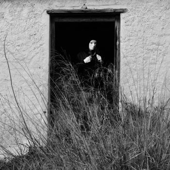 Masked figure by broken door of abandoned house obscured by overgrown plants.