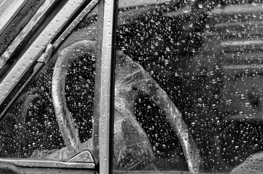 Vintage car interior through rain covered window steering wheel wrapped in plastic. Black and white.