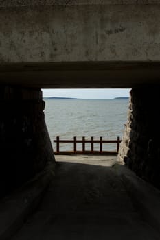 A tunnel opening with a rusty metal barrier and the sea beyond on a sunny day with cloud and an island in the distance.