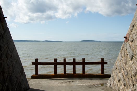 A tunnel opening with a rusty metal barrier and the sea beyond on a sunny day with cloud and an island in the distance.