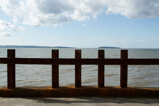 A rusty metal barrier provides security against the sea on a sunny day with cloud and an island in the distance.