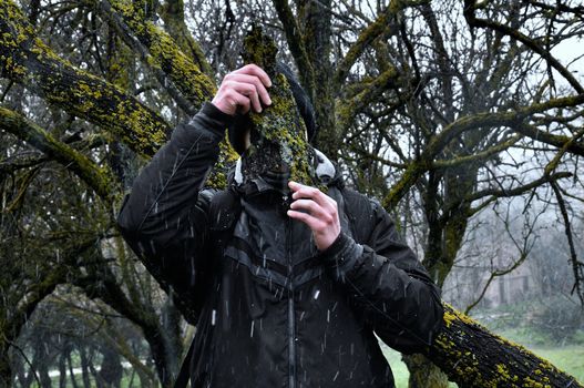 Man with bark peel mask under tree branches and falling snow in the woods.