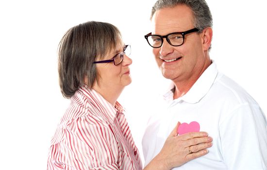 Senior couple with pink heart isolated on white. Woman placing paper heart on chest of her man