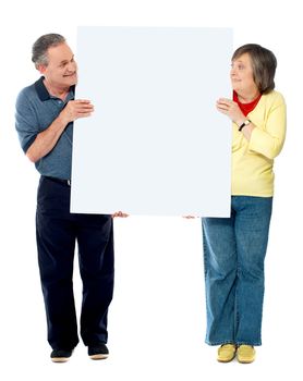 Mature couple presenting blank white billboard, looking at each other