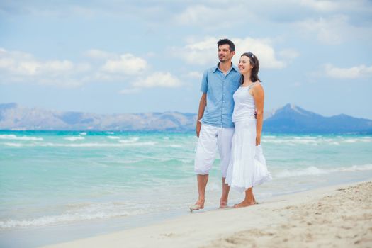 Back view of happy young couple walking on a deserted tropical beach with bright clear blue sky