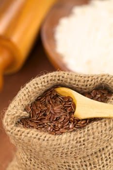 Brown flax seeds in jute bag with flour and rolling pin in the back (Selective Focus, Focus on the front flax seeds on the wooden spoon)