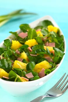 Fresh watercress, pineapple and ham salad in elongate white bowl with a fork on the side (Selective Focus, Focus on the two pineapple pieces one third into the salad)