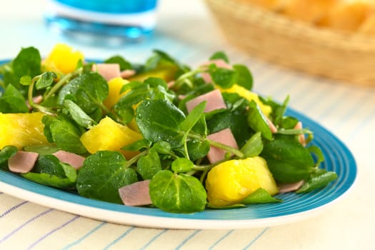 Fresh watercress, pineapple and ham salad on blue plate with bread basket and glass in the back (Selective Focus, Focus on the pineapple pieces in the front)