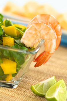 Cooked shrimps on the rim of a glass bowl with fresh watercress, mango and avocado salad with lime wedges on the side and baguette in the back (Selective Focus, Focus on the first shrimp)