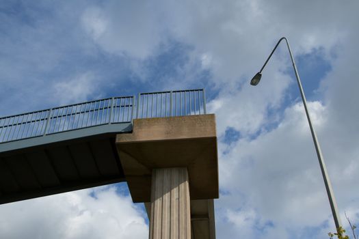 A raised walkway with a concrete supporting pillar and metal hand railings and a street light post against a blue cloudy sky.