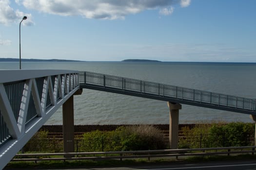 A metal footbridge leads to a ramp supported by concrete pillars with the sea, island, blue sky and cloud in the background.