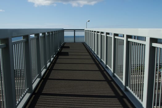 A metal footbridge with sakety railings over a road with the sea and blue sky in the background.