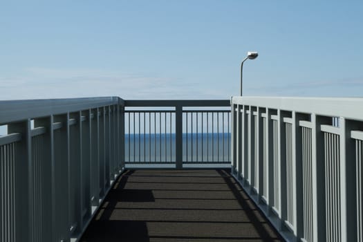 A metal footbridge with sakety railings over a road with the sea and blue sky in the background.