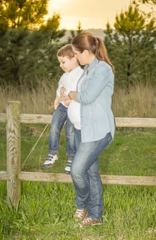 Pregnant mother sitting her cute son on a wooden fence in a field at sunset