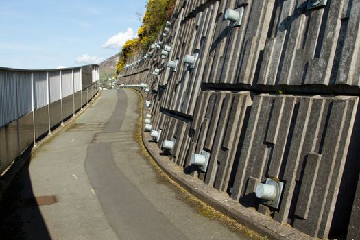 A footpath with railings winds along next to an engineered retaining wall with metal pins against a blue sky.