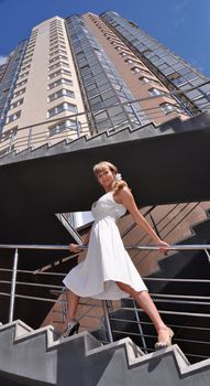 Portrait of a young pretty woman in white dress walking up the stairs on the background of high building and blue sky