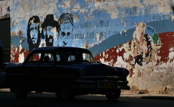 American taxi car on the street in Havana, Cuba. Behind a car painted wall with images of Che Gevarra,Fidel Castro and propaganda words